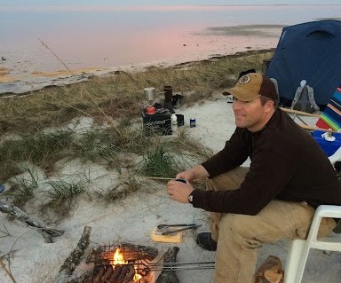 A person sits on the beach looking out over a campfire and the water.